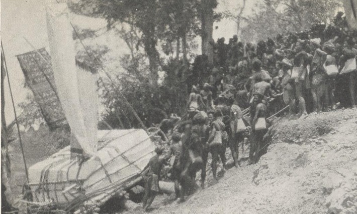 Crowd dragging a megalithic tombstone on a wooden sledge, with a large ikat textile flown as a banner on a pole