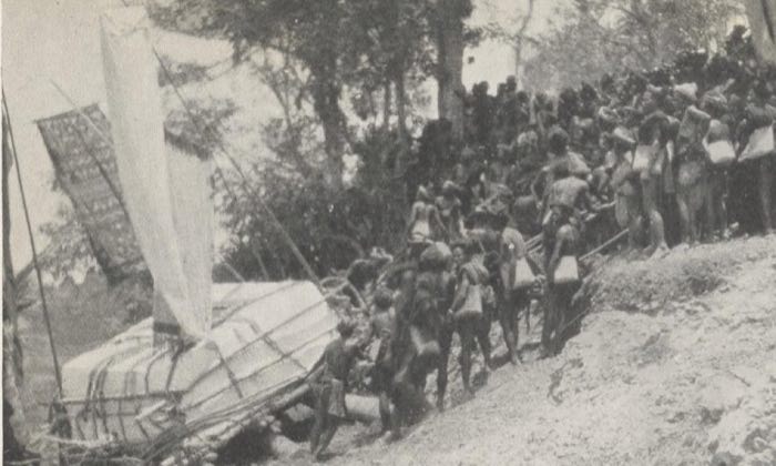Crowd dragging a megalithic tombstone on a wooden sledge, with a large ikat textile flown as a banner on a pole